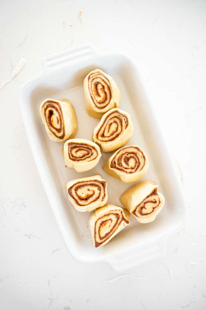 cut cinnamon roll dough in a white baking dish on a white marble counter.