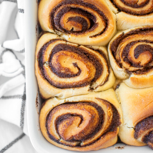 golden brown homemade cinnamon rolls in a white baking dish on a white marble counter.