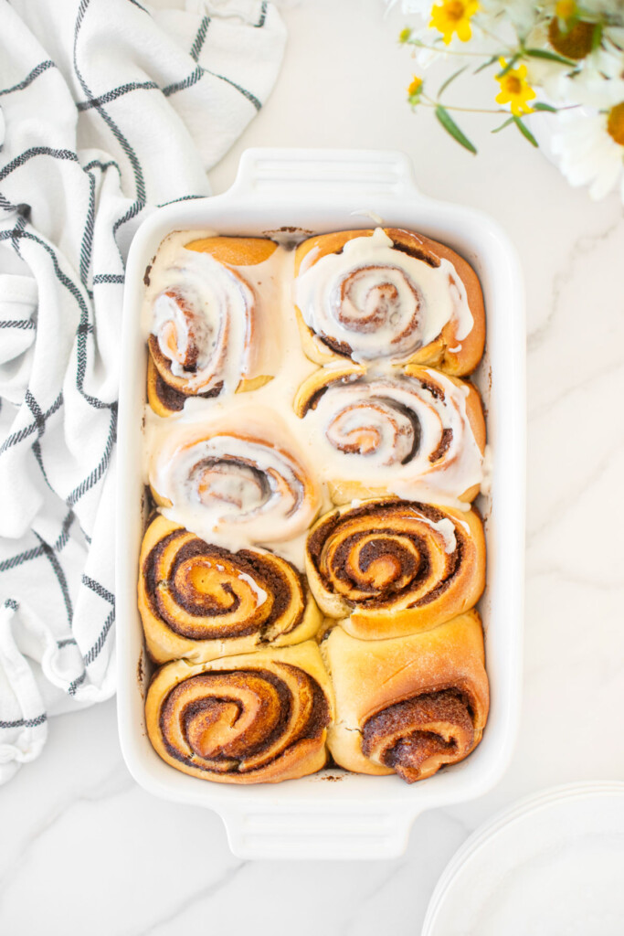 homemade cinnamon rolls in a white ceramic baking dish partially frosted on a white marble counter with fresh flowers.