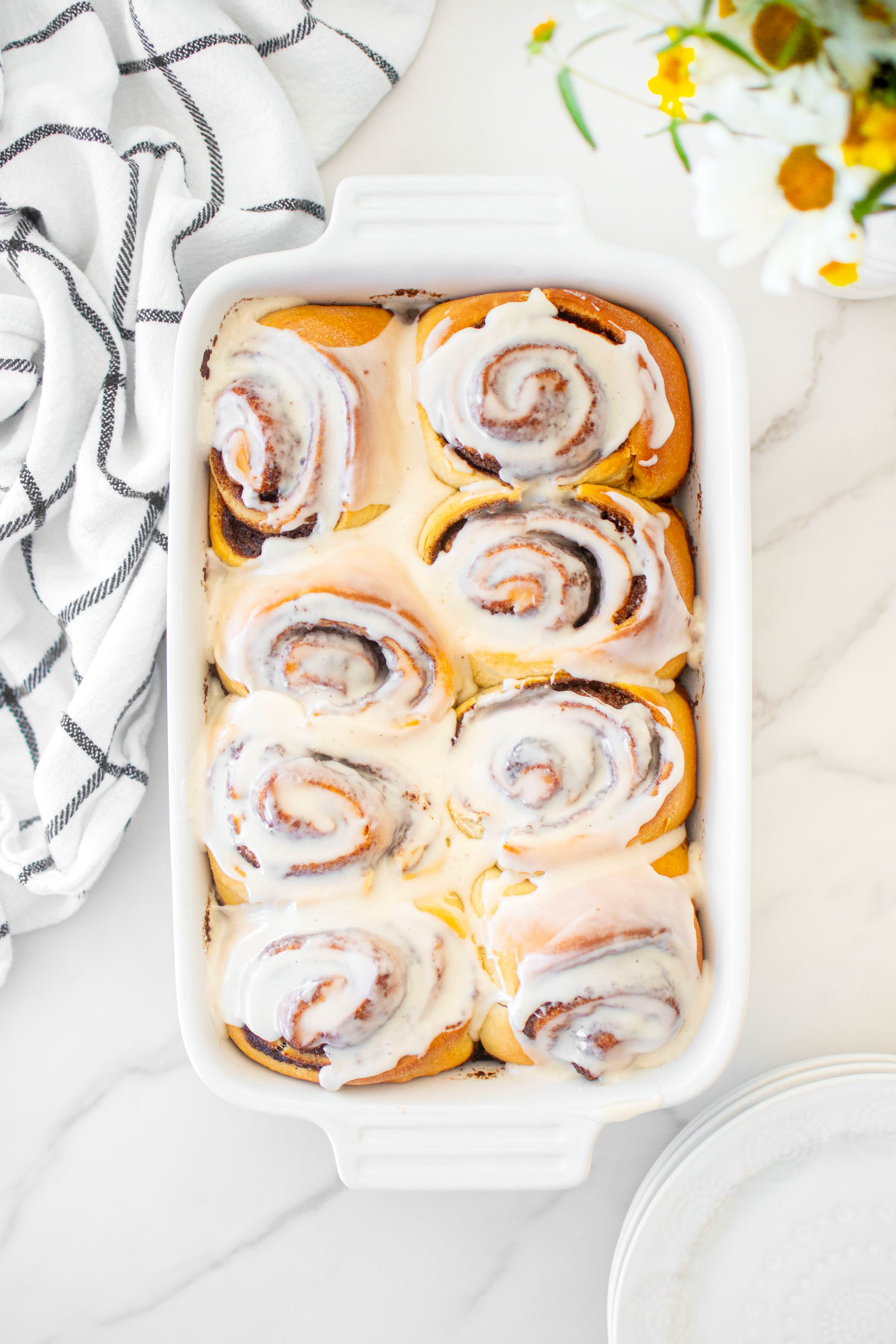 homemade cinnamon rolls in a white ceramic baking dish frosted on a white marble counter with fresh flowers.