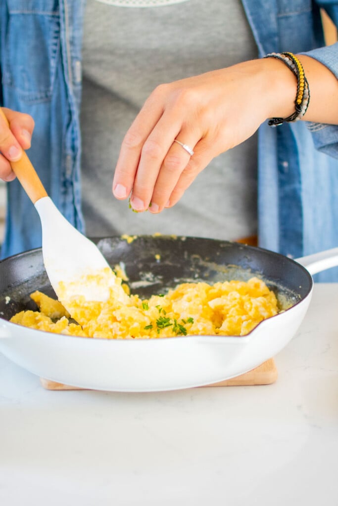 woman adding fresh chives to fluffy scrambled eggs with sour cream in a white cast iron skillet at the kitchen counter.