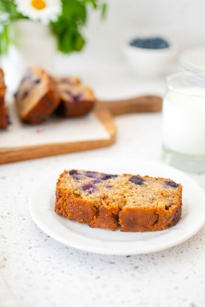 slice of blueberry buttermilk banana bread on a white plate with a glass of milkk.