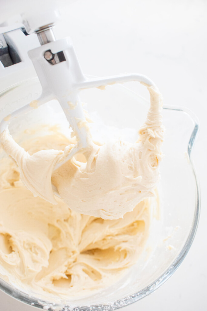 cream cheese frosting in a glass stand mixing bowl on a white marble counter.