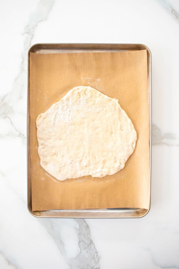 chicago style thin crust pizza dough on a baking sheet on a white marblec ounter.