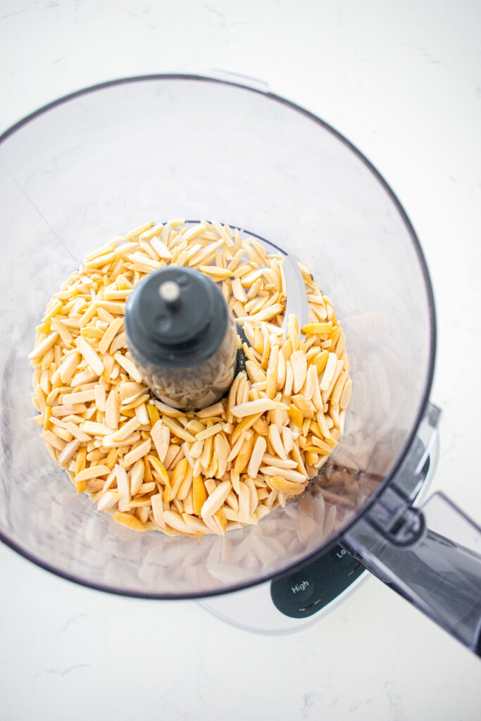 food processor with blanched slivered almonds on a marble counter.