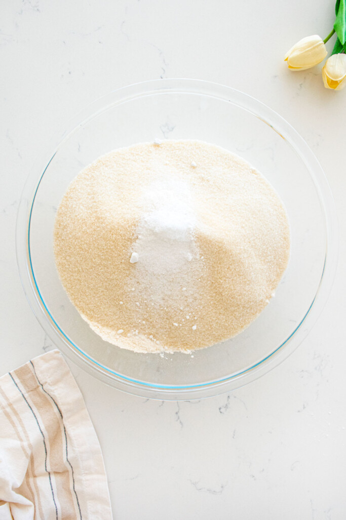 large glass mixing bowl with cake flour, cane sugar, baking powder and salt on a marble counter.