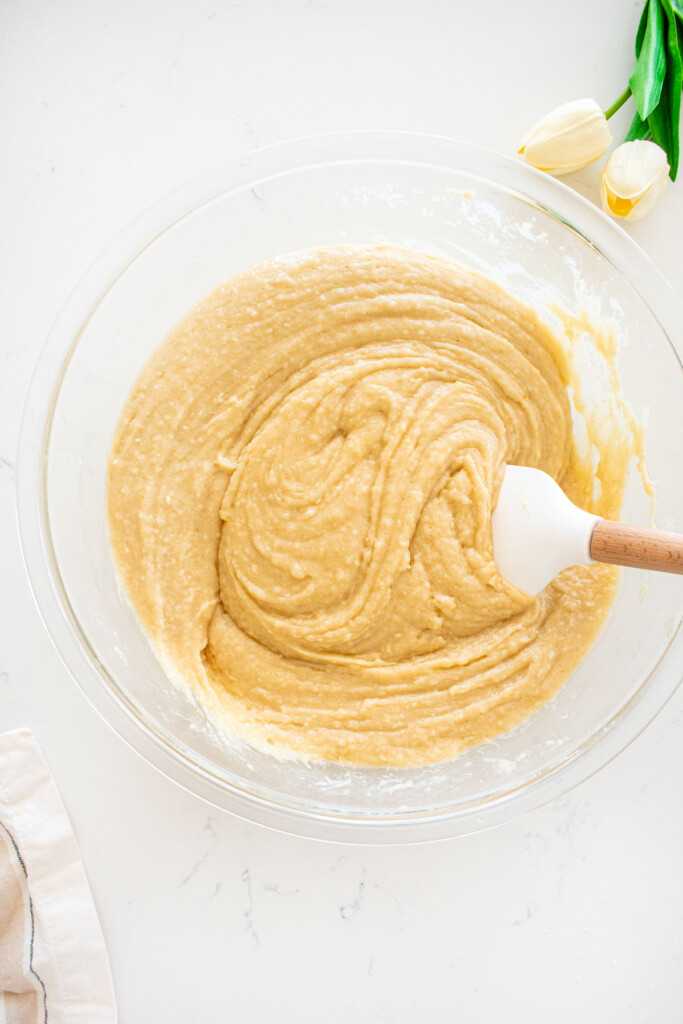 eggless coconut cake batter mixed together in a glass mixing bowl with a spatula on a marble counter.