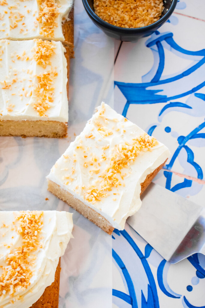 square slice of eggless coconut sheet cake on a cake spatula on a blue tile table.