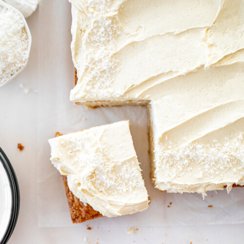 eggless coconut sheet cake with coconut buttercream frosting on a marble counter with one slice cut out of it.