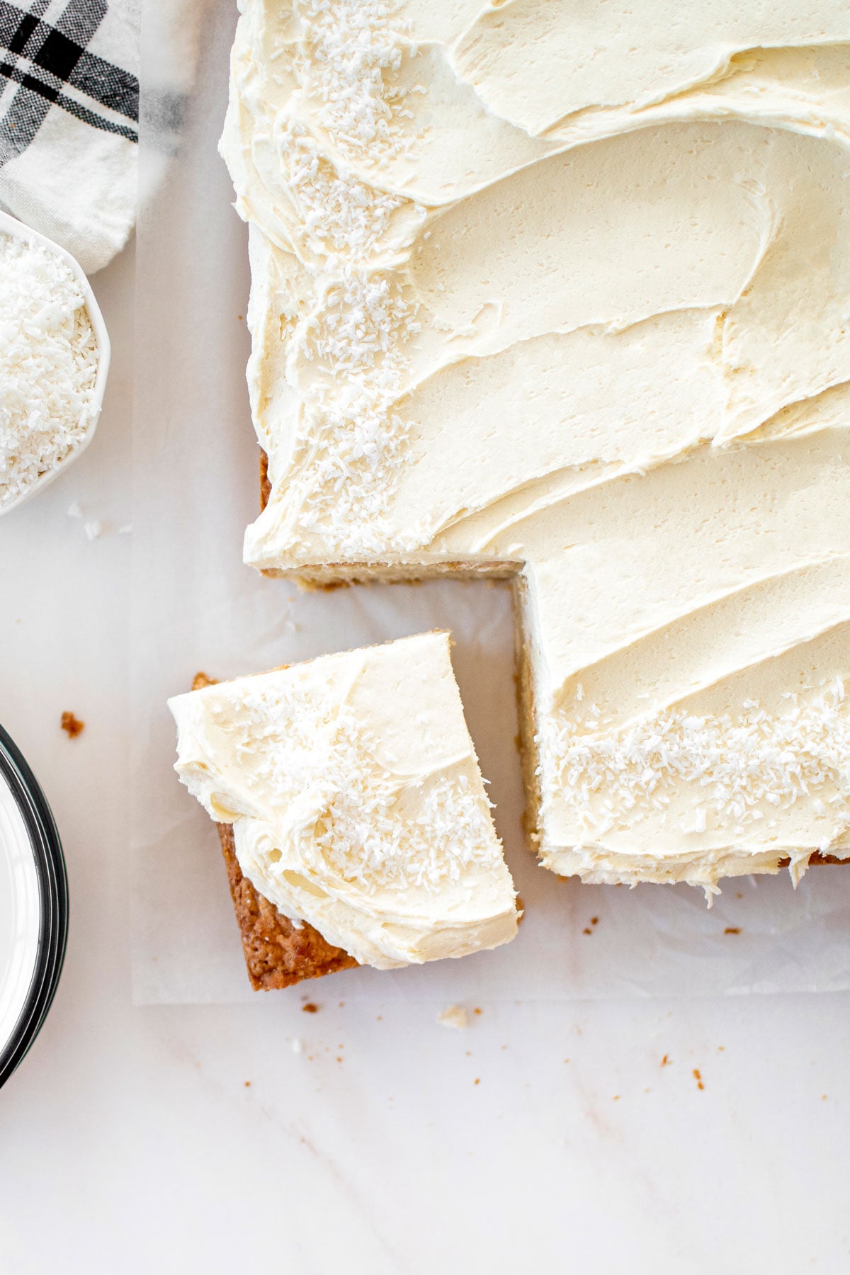 eggless coconut sheet cake with coconut buttercream frosting on a marble counter with one slice cut out of it.