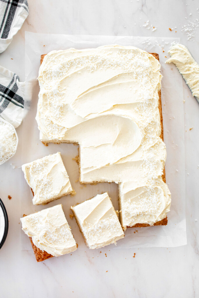eggless coconut sheet cake with coconut buttercream frosting on a marble counter with three slices cut out of it.