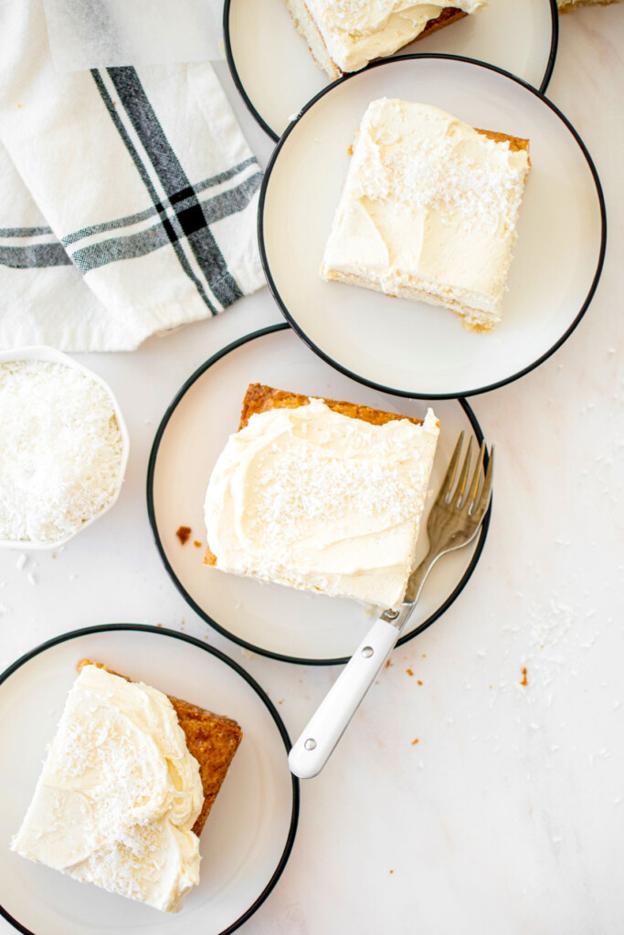 square slices of eggless coconut sheet cake with coconut buttercream on cake plates on a marble counter.