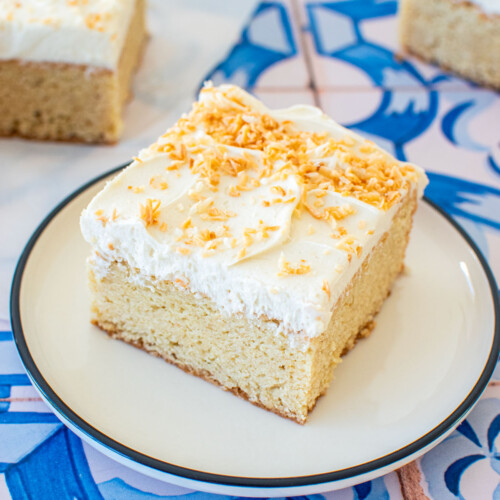 square slice of eggless coconut sheet cake with coconut buttercream and toasted coconut on a plate on a blue tile table.