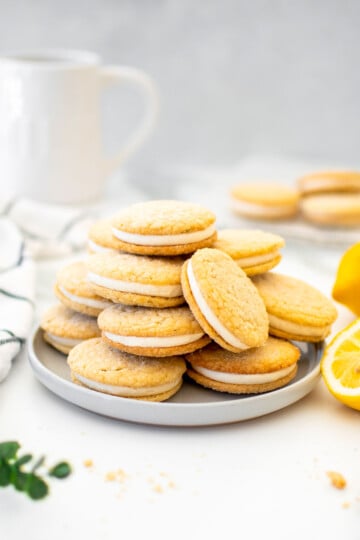 lemon rosemary sandwich cookies on a plate on a marble counter with fresh lemons and a pitcher of milk.