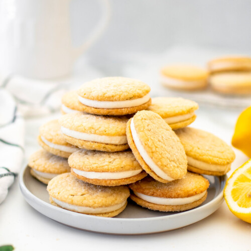 lemon rosemary sandwich cookies on a plate on a marble counter with fresh lemons and a pitcher of milk.