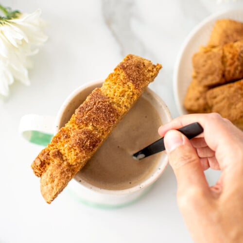 snickerdoodle biscotti sitting on top of a mug of coffee that a woman is stirring on a white marble counter.