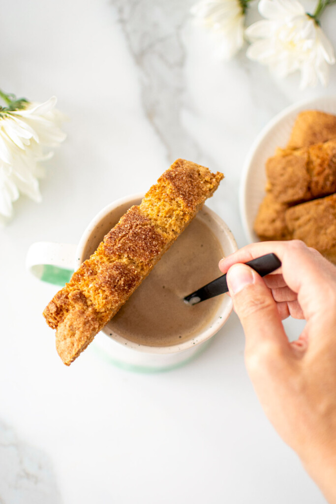 snickerdoodle biscotti sitting on top of a mug of coffee that a woman is stirring on a white marble counter.