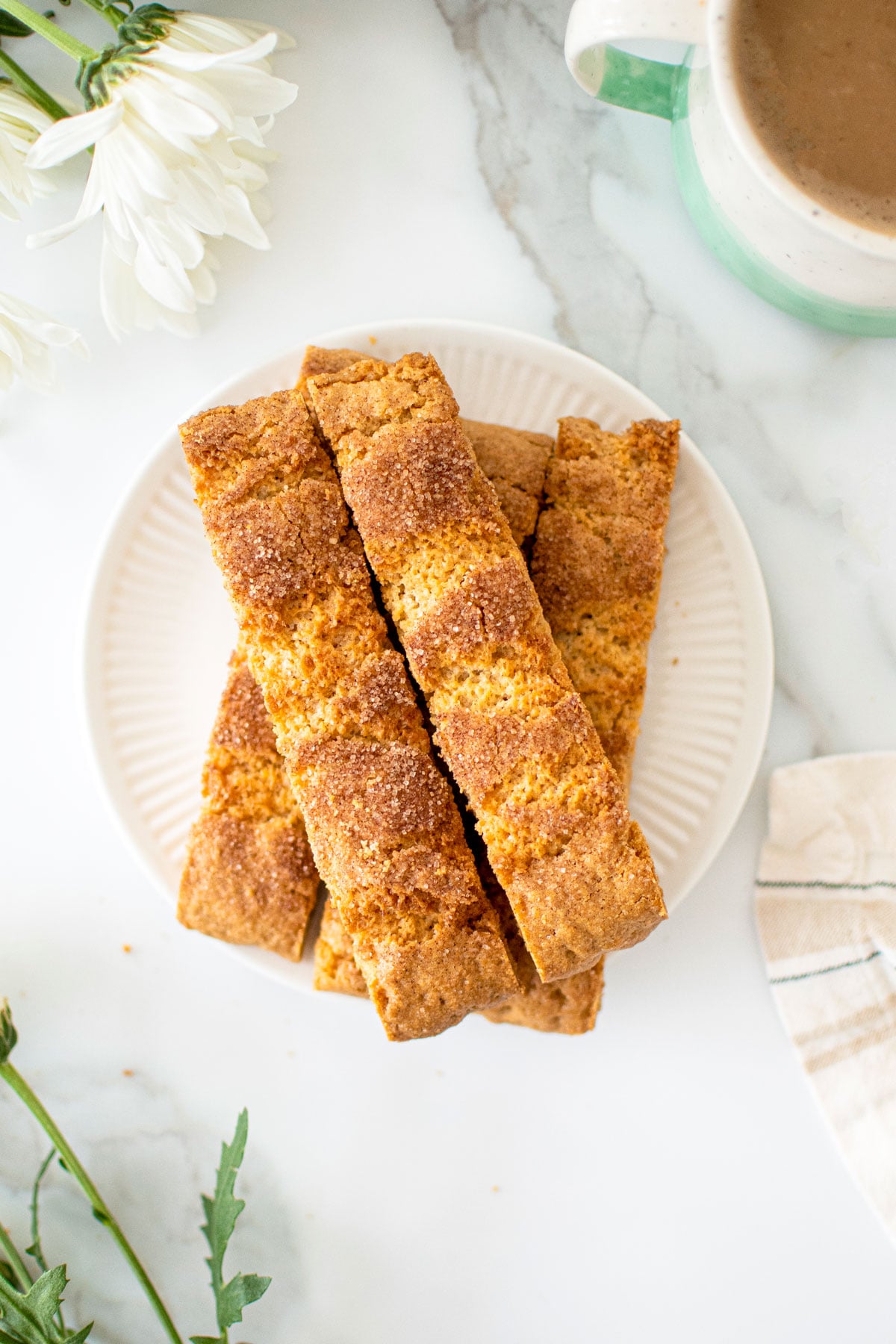 snickerdoodle biscotti on a plate on a white marble counter with fresh flowers and coffee.