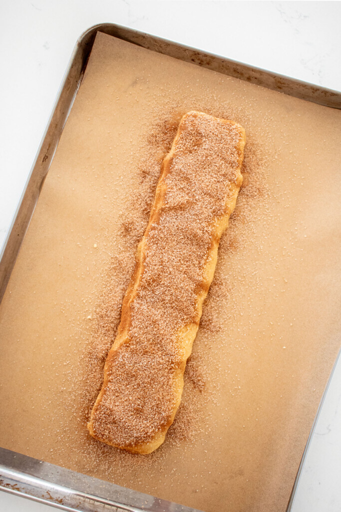 snickerdoodle biscotti cookie dough log with cinnamon and sugar on top on a baking sheet on a white marble counter.
