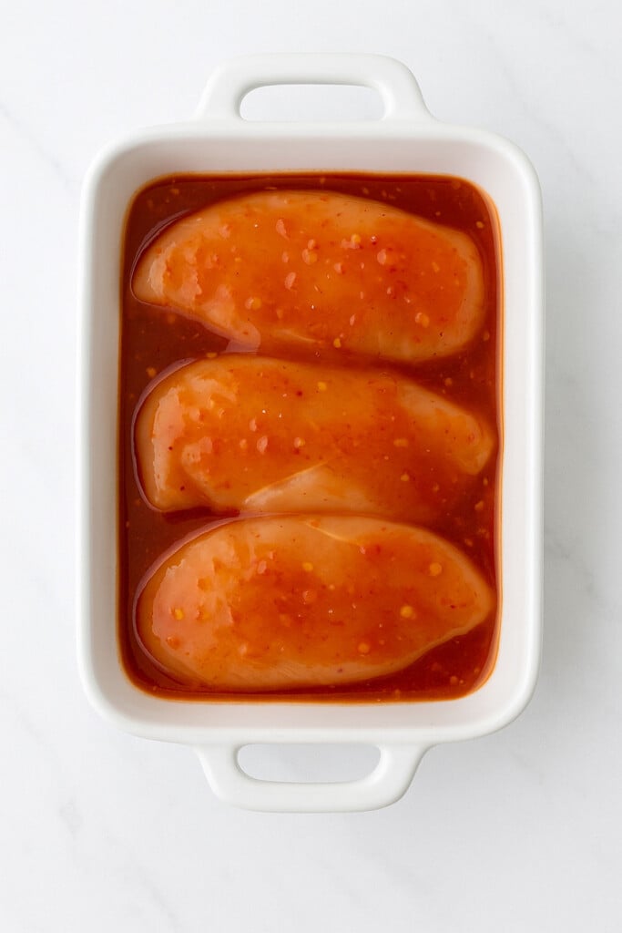 chicken breasts marinating in sweet chili sauce in a white baking dish on a marble countertop.