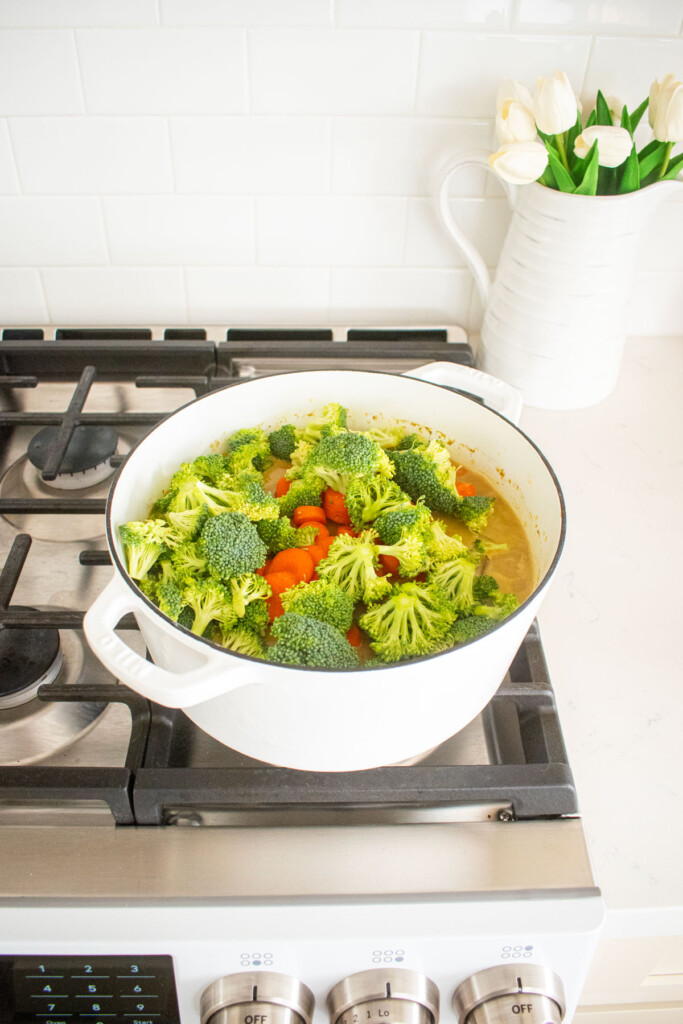 yellow curry chicken with bone broth with added broccoli and carrots in a white dutch oven on a stovetop.