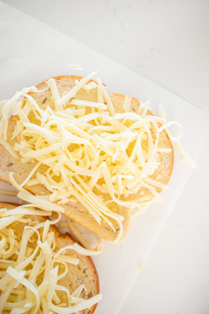 sourdough bread slices with melted butter, garlic and shredded mozzarella on parchment paper on a marble counter.