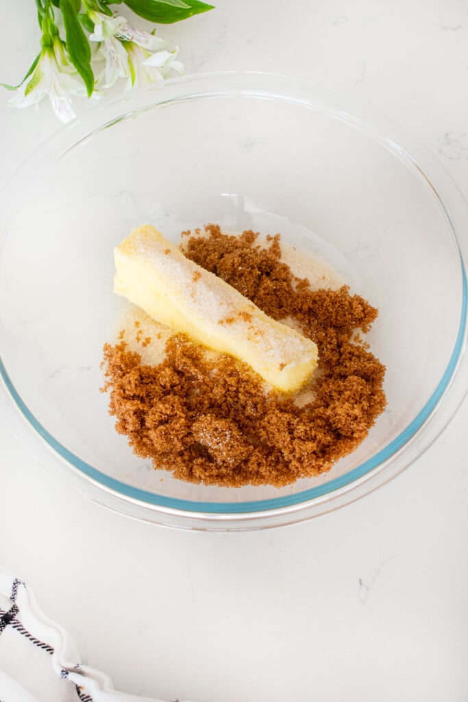 glass mixing bowl with butter, cane sugar and dark brown sugar on a white marble counter.