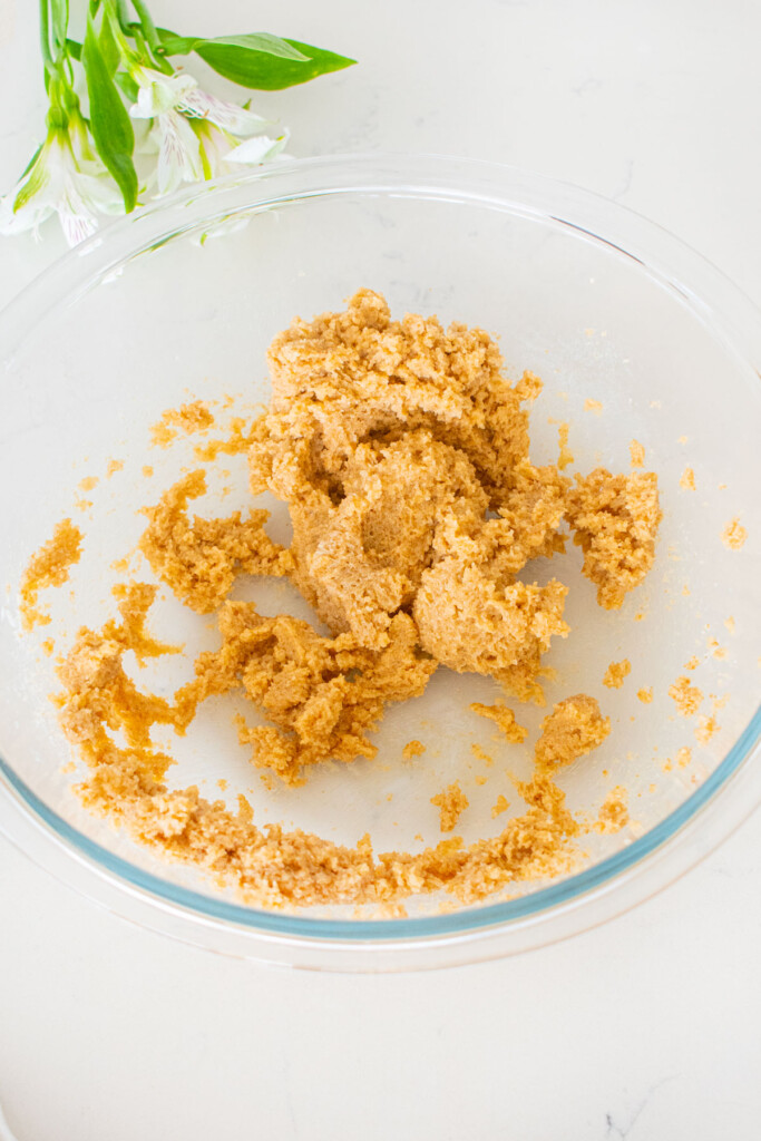 glass mixing bowl with creamed butter, cane sugar and dark brown sugar on a white marble counter.
