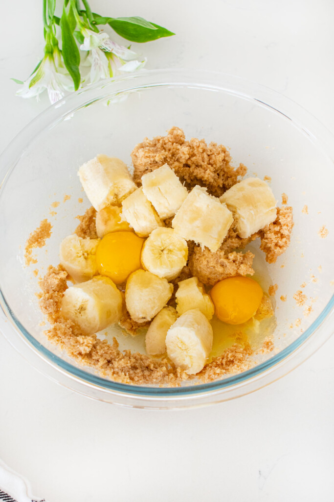 glass mixing bowl with creamed butter, cane sugar and dark brown sugar with banana chunks and eggs on top on a white marble counter.
