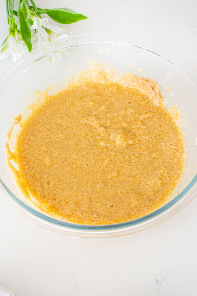 wet ingredients for banana bread cake mixed together in a glass mixing bowl on a white marble counter.