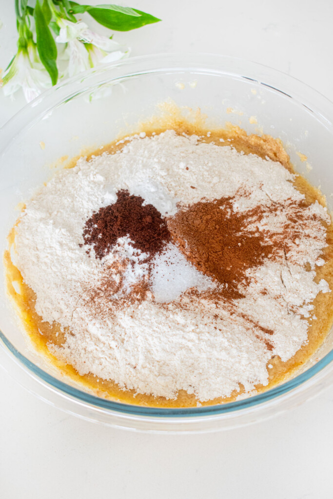dry ingredients added into wet ingredients for banana bread cake in a glass mixing bowl on a white marble counter.