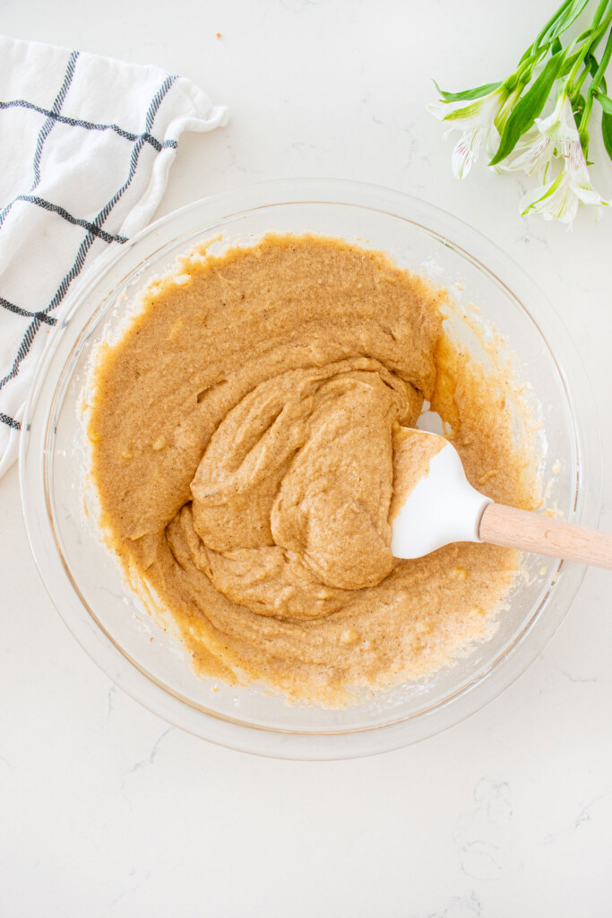 glass mixing bowl with banana bread cake batter and a spatula on a white marble counter.