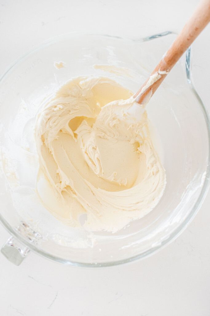 creamy buttercream frosting in a glass mixing bowl with a spatula on a white marble counter.
