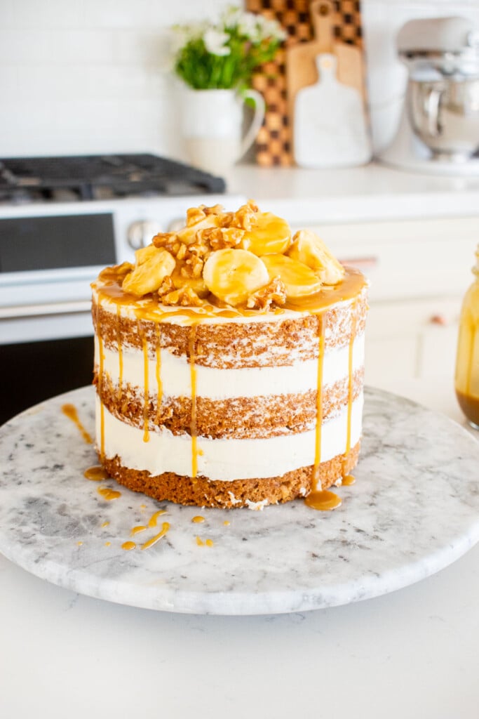 banana bread semi naked cake with fresh banana slices, walnuts and butterscotch sauce on a marble turntable on a white kitchen counter.