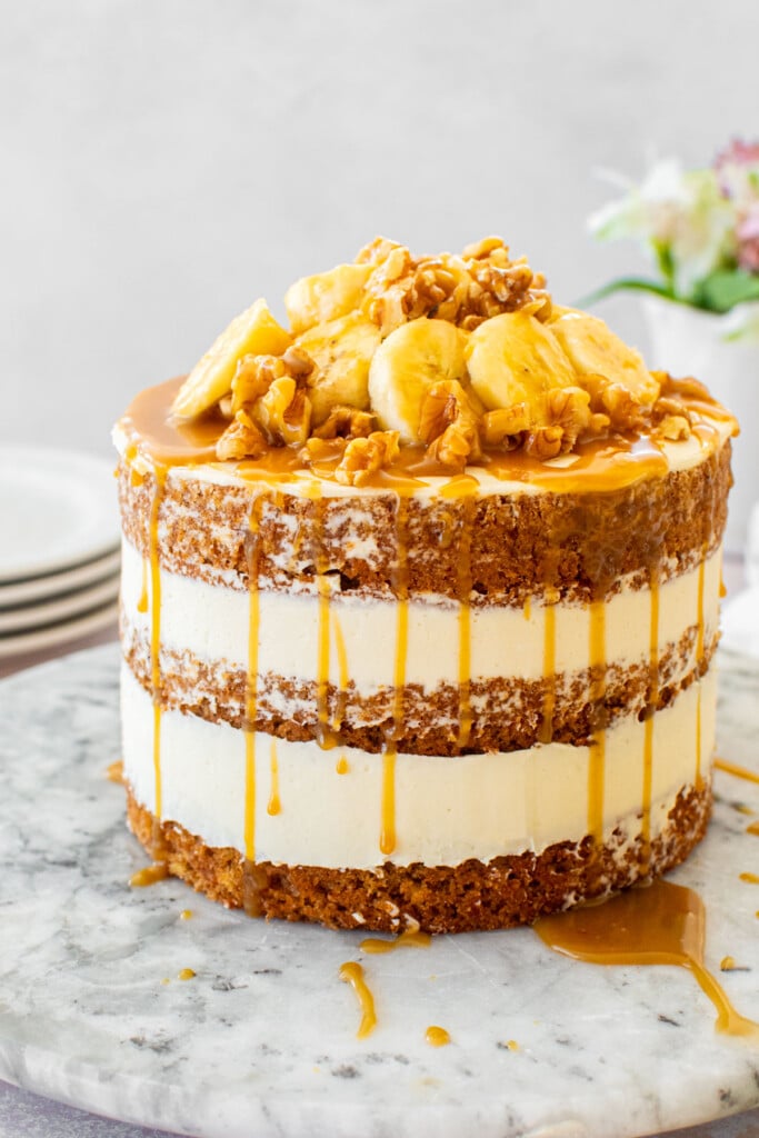 banana bread semi naked cake with fresh banana slices, walnuts and butterscotch sauce on a marble serving plate on a table with fresh flowers.