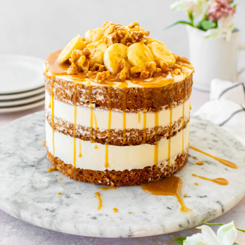 banana bread semi naked cake with fresh banana slices, walnuts and butterscotch sauce on a marble serving plate on a table with fresh flowers.