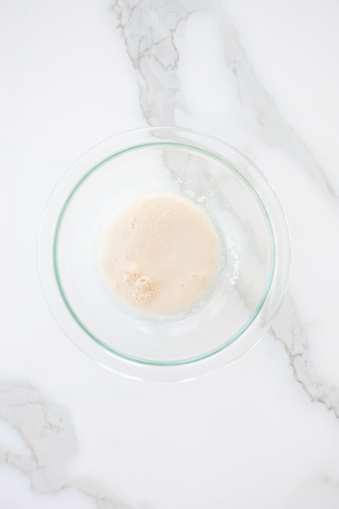 yeast and water in a glass mixing bowl on a white marble counter.