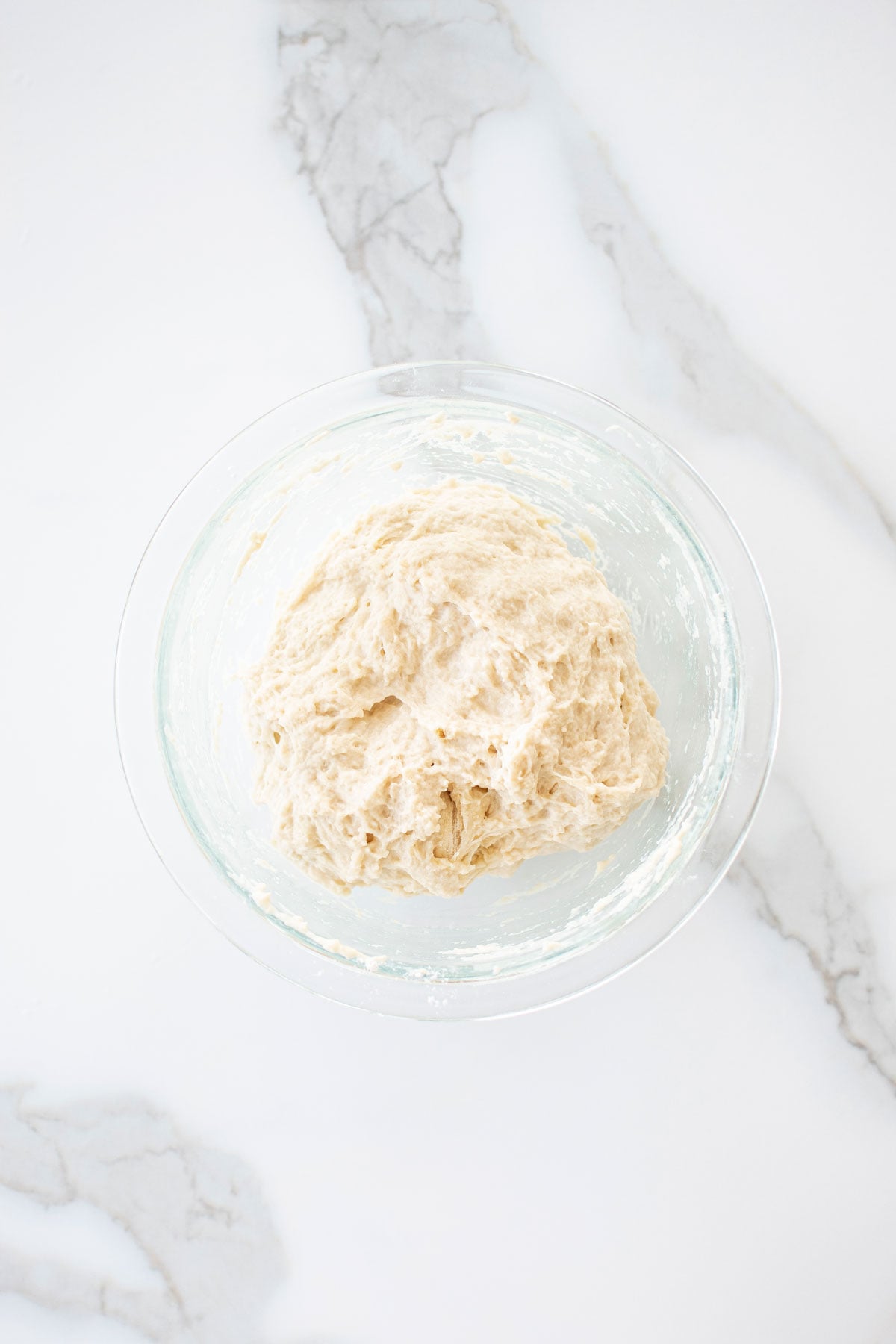 chicago style thin crust pizza dough in a glass mixing bowl on a white marble counter.