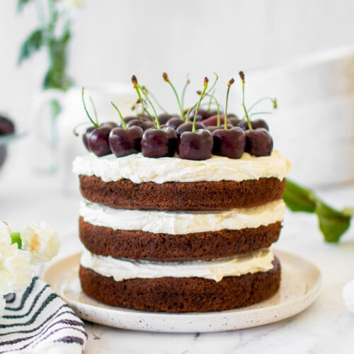 chocolate cherry whipped cream cake with fresh cherries on top on a cake plate on a marble counter with fresh flowers.