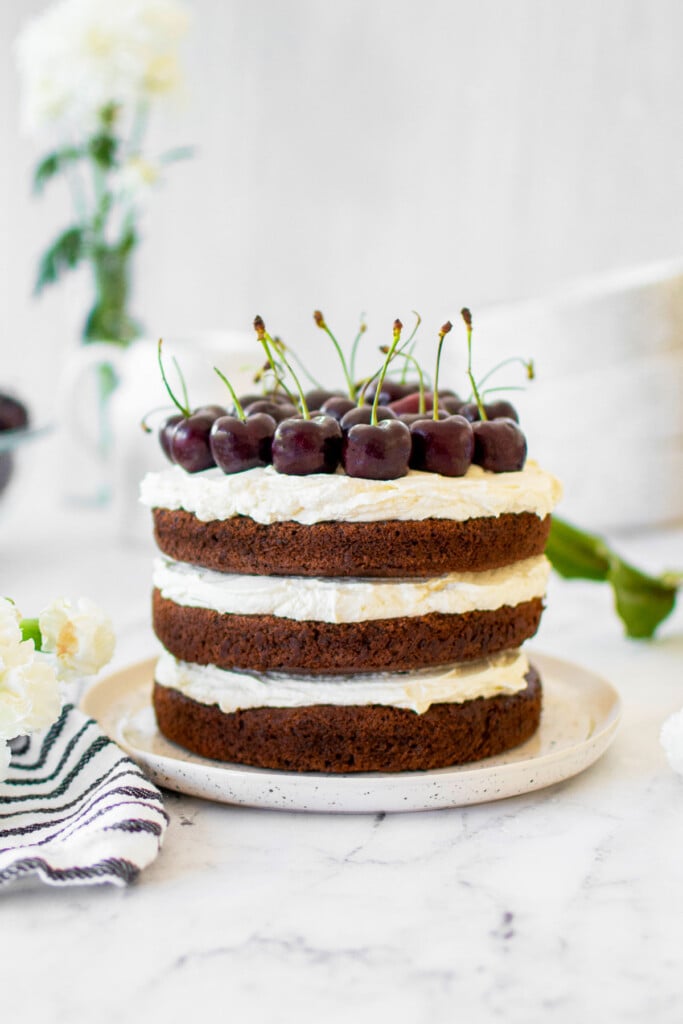 chocolate cherry whipped cream cake with fresh cherries on top on a cake plate on a marble counter with fresh flowers.