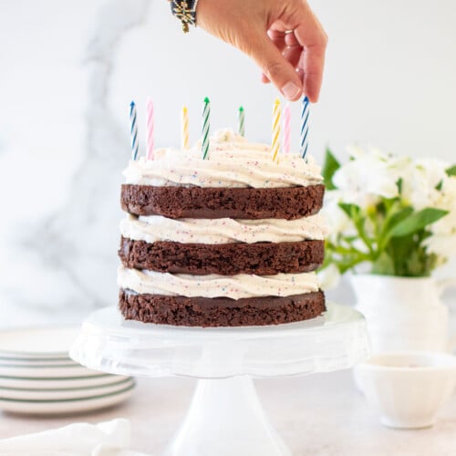 woman placing a candle in an eggless chocolate birthday cake with cream cheese funfetti frosting on a cake stand on a table with fresh flowers.