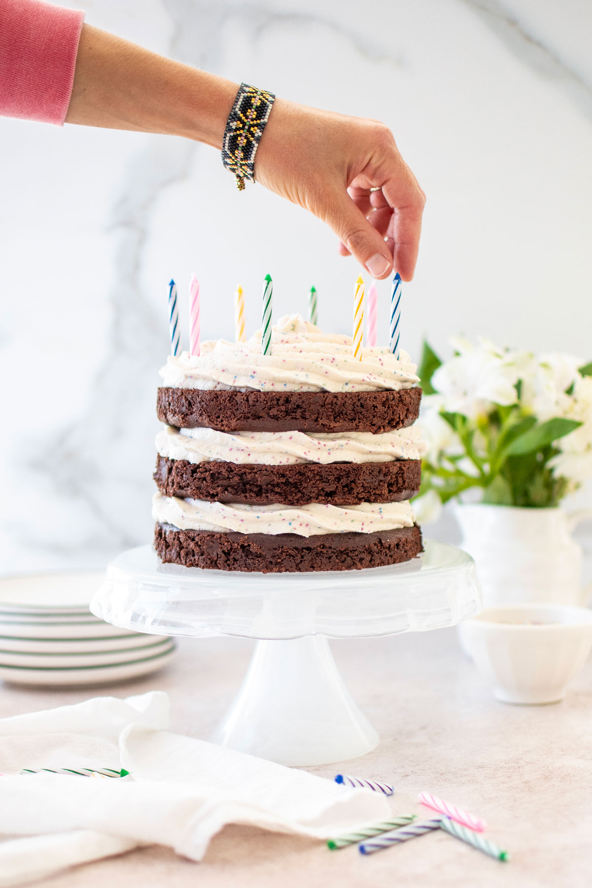 woman placing a candle in an eggless chocolate birthday cake with cream cheese funfetti frosting on a cake stand on a table with fresh flowers.