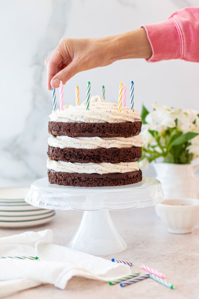 woman placing a candle in an eggless chocolate birthday cake with cream cheese funfetti frosting on a cake stand on a table with fresh flowers.