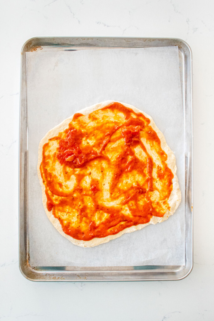 pizza dough on a baking sheet with tomato sauce on it on a white marble counter.