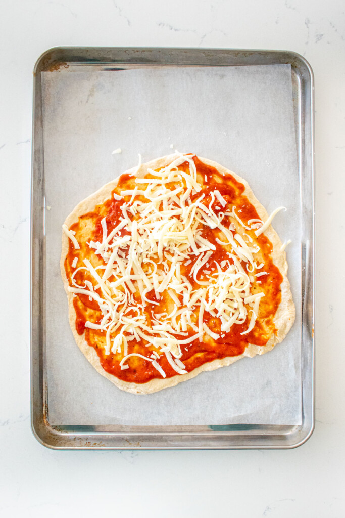 pizza dough with tomato sauce and cheese on a baking sheet on a white marble counter.