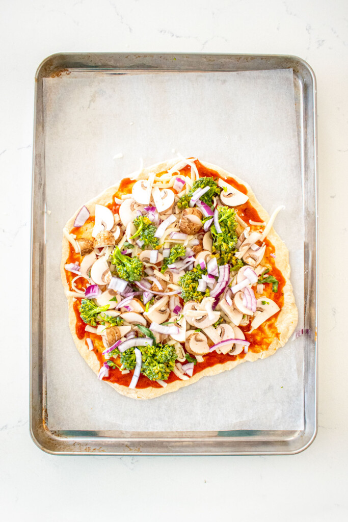 pizza dough with tomato sauce, cheese and veggies on a baking sheet on a white marble counter.