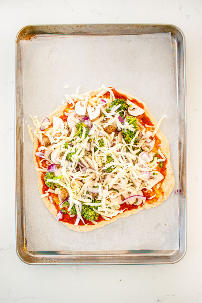 pizza dough with tomato sauce, cheese, and veggies on a baking sheet on a white marble counter.