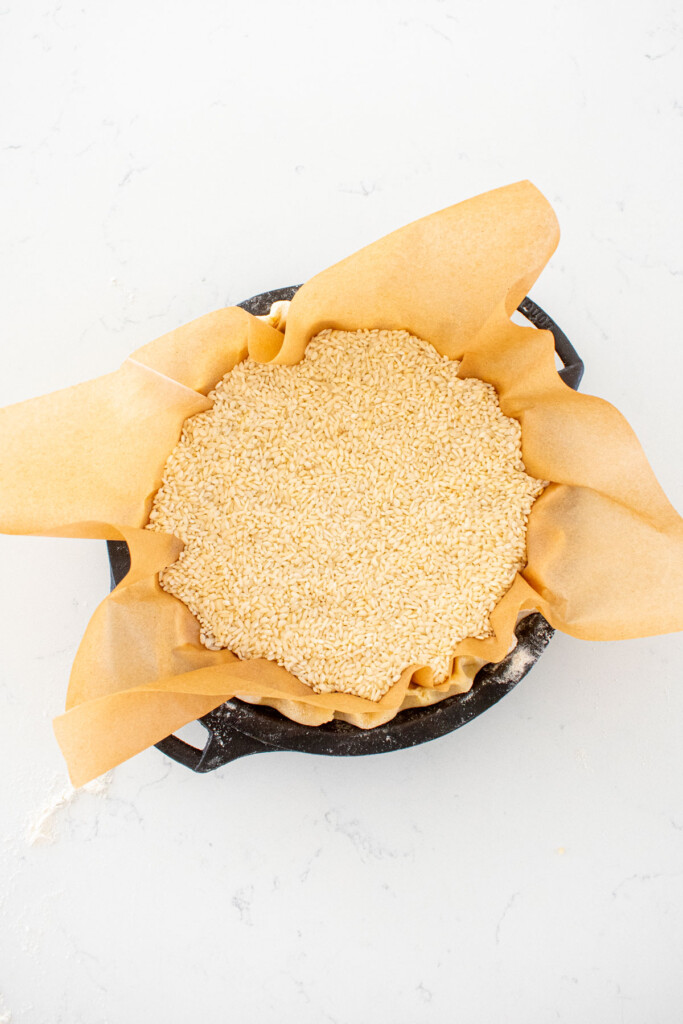 pie crust in a cast iron pie pan with parchment paper and rice inside on a white marble counter.