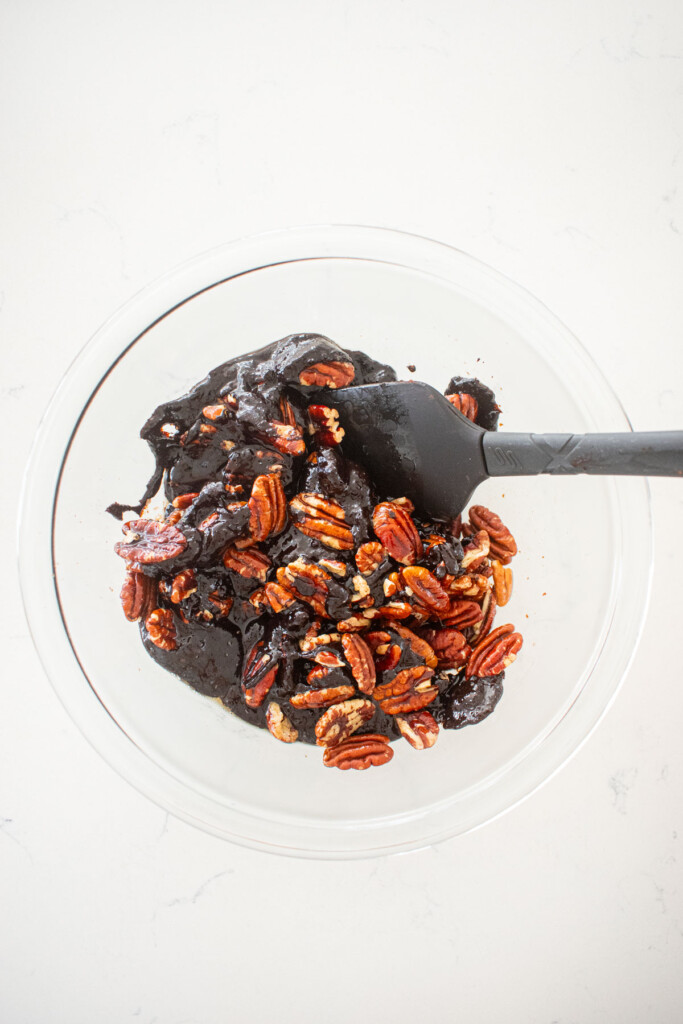 chocolate bourbon pecan pie filling with pecans being stirred in a glass mixing bowl on a white marble counter.