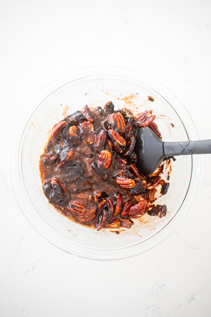 chocolate bourbon pecan pie filling with pecans being stirred in a glass mixing bowl on a white marble counter.